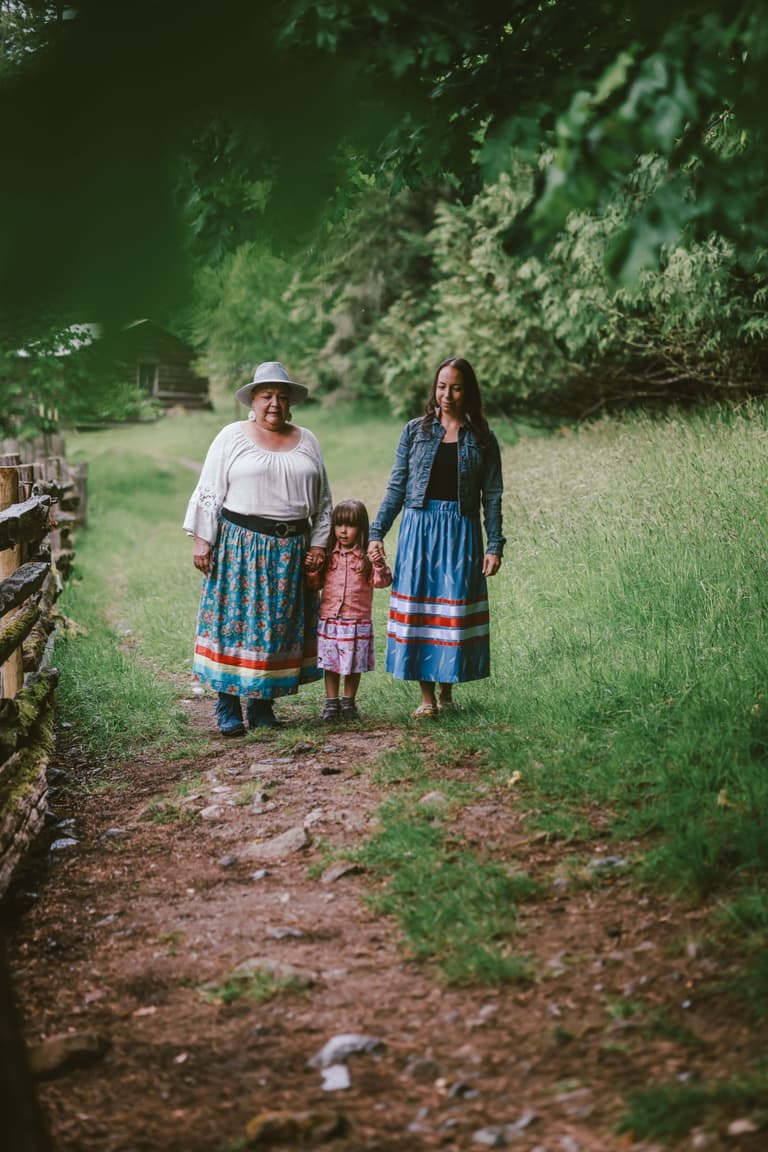 Two women and a child in ribbon skirts on a wet orchard path