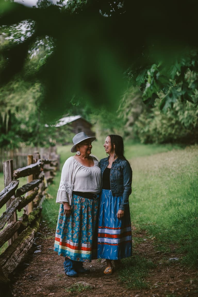 Two women in ribbon skirts standing together beside a split rail fence