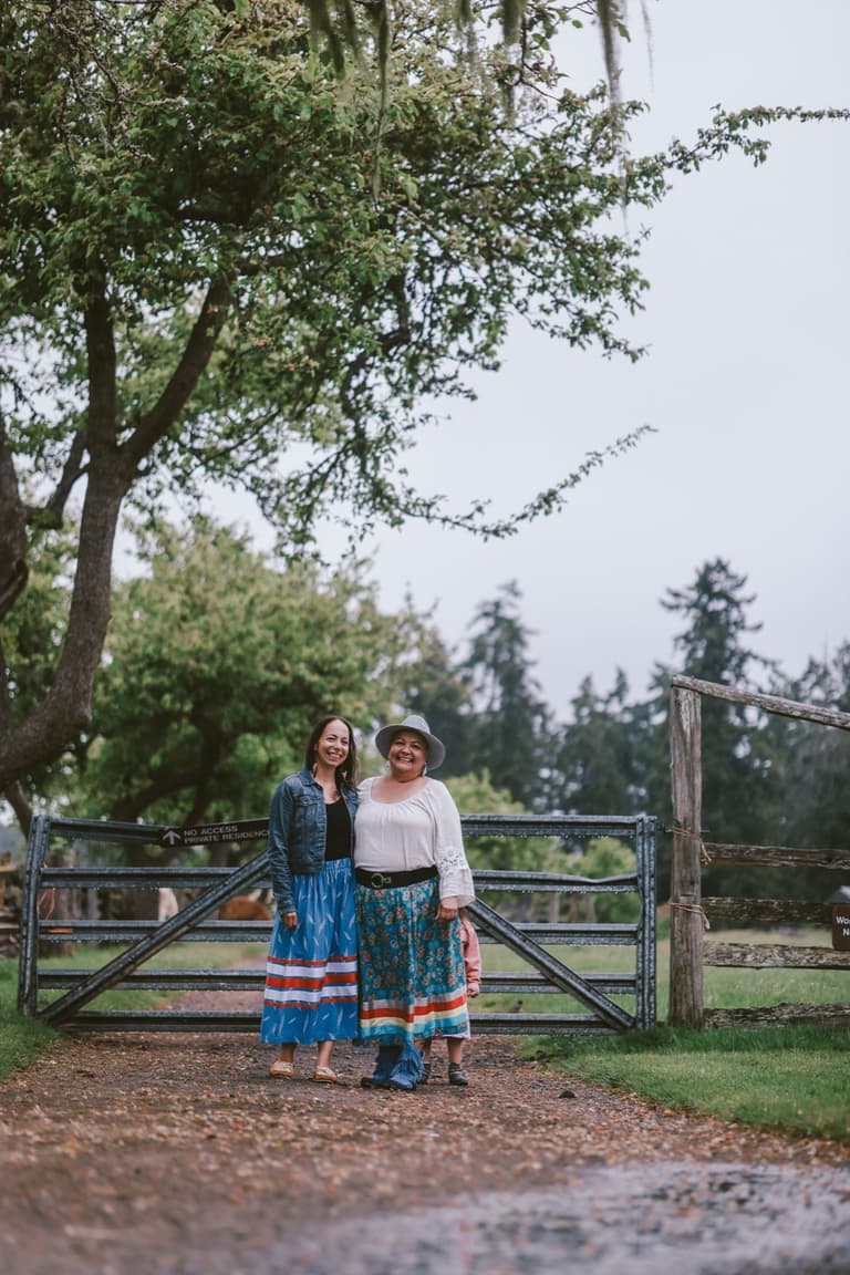 Two women in ribbon skirts at a farm gate